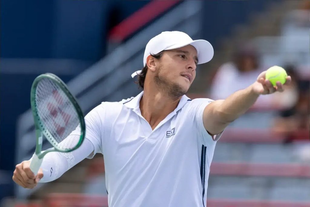 Tennis player in white cap and EA7 polo holding a green racket and a yellow ball, preparing to serve at the US Open
