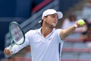 Tennis player in white cap and EA7 polo holding a green racket and a yellow ball, preparing to serve at the US Open