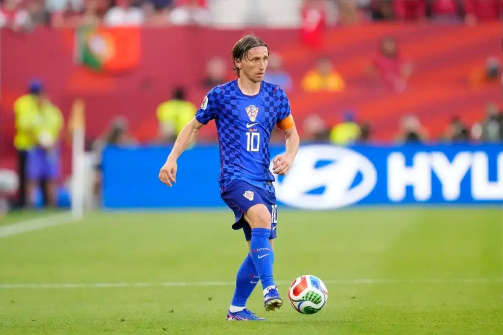Player in blue checkered Croatia kit, number 10 and captain's armband, dribbling the ball on a green pitch during a match.