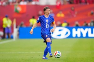 Player in blue checkered Croatia kit, number 10 and captain's armband, dribbling the ball on a green pitch during a match.