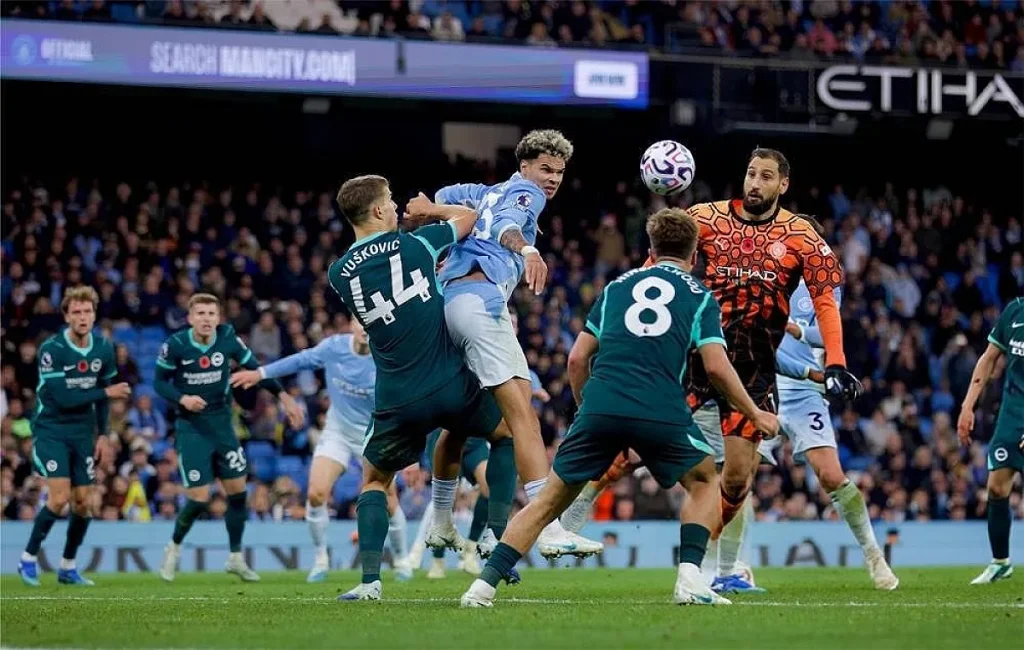 Jogadores do Brighton de verde e do Man City de azul-claro disputam uma bola no ar em frente à baliza no Etihad, com Vušković (44) em destaque.