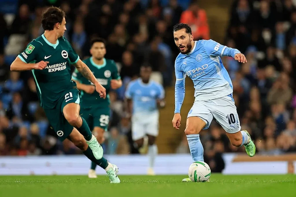 Jogador do Man City, de camisola azul-clara e número 10, conduz a bola em frente a adversário do Brighton, em jogo da Taça da Liga.