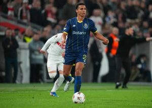 Player in navy Porto kit with number 25 dribbling the ball on the pitch, Europa League match, opponent in white chasing behind.