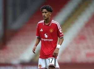 Jovem de caracóis escuros com camisola vermelha do Manchester United, número 17, em campo com bancadas desfocadas ao fundo.