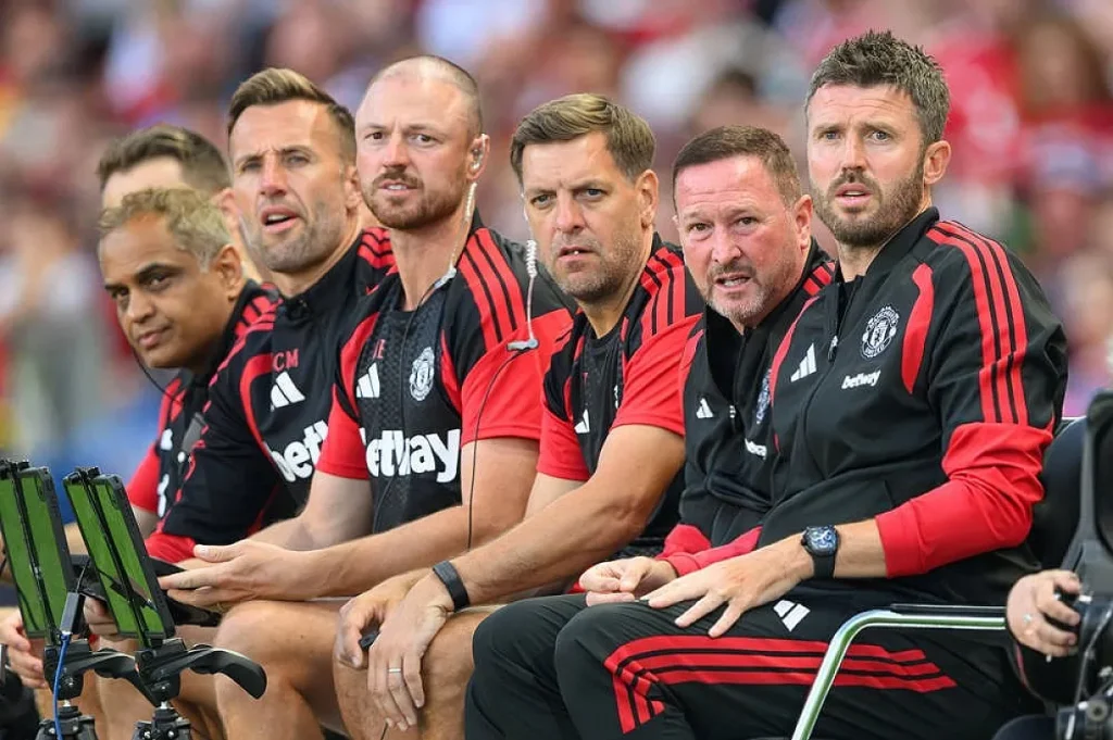 Equipa técnica do Manchester United no banco, de fato de treino preto e vermelho, com patrocínio Betway, a acompanhar um jogo.