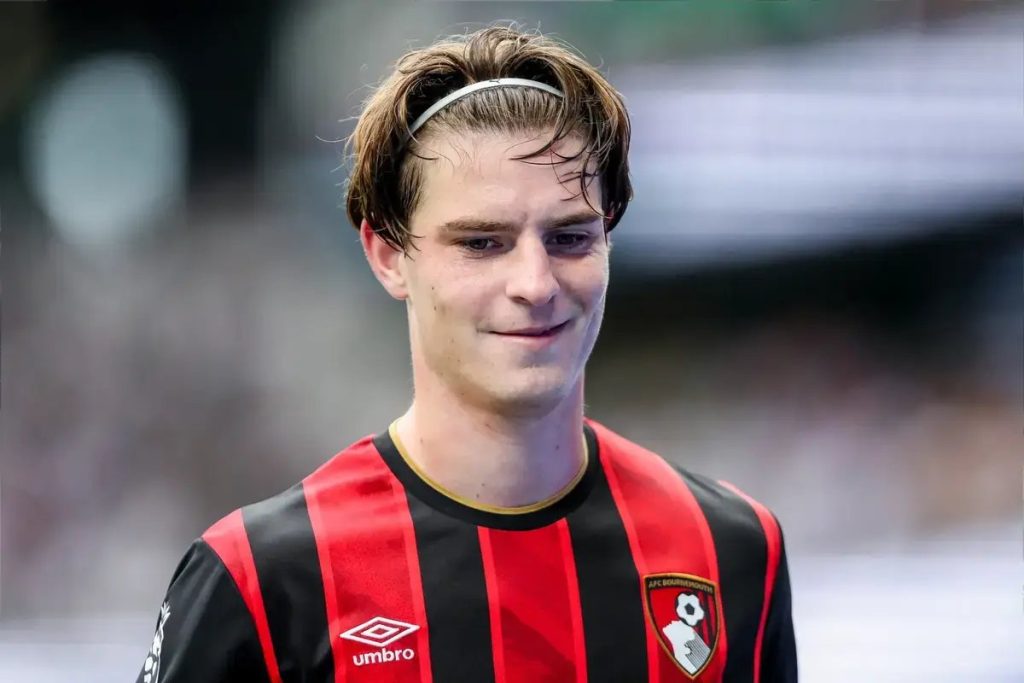 Young man with tousled brown hair and a white headband, smiling, wearing an AFC Bournemouth red-and-black striped kit with Umbro logo at a stadium.