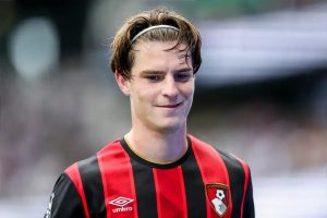 Young man with tousled brown hair and a white headband, smiling, wearing an AFC Bournemouth red-and-black striped kit with Umbro logo at a stadium.