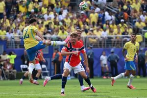 Brazil player in yellow kit with number 20 kicks the ball in mid-air while a Norway player in red and white defends, in a stadium.