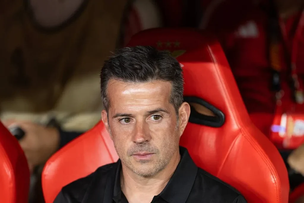 Treinador de bancada vestido de preto, sentado em cadeira vermelha num estádio, com equipamento vermelho em segundo plano