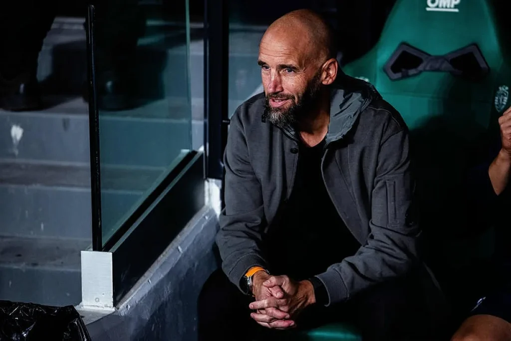 Homem careca e barbudo, de casaco cinzento, sentado num banco de suplentes verde com um relógio laranja, em estádio à noite.