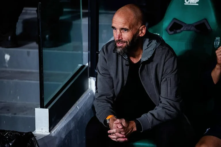 Homem careca e barbudo, de casaco cinzento, sentado num banco de suplentes verde com um relógio laranja, em estádio à noite.