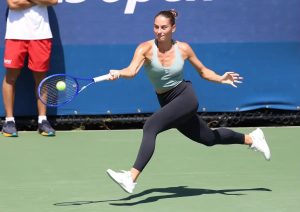 A tennis player in a teal top and black leggings sprints to hit a forehand on a green court, with a "US Open" backdrop behind her.