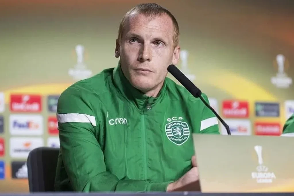 Homem de t-shirt verde do Sporting CP numa conferência de imprensa da Liga Europa, com microfone à frente.