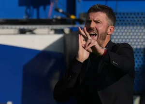 Treinador de fato escuro a gesticular e gritar à beira do relvado, com fundo azul de estádio. Contexto: artigo sobre Michael Carrick e o Manchester United.