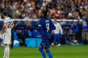 Michael Olise in a blue France jersey, number 11, gesturing during a match as a player in white looks on at the pitch