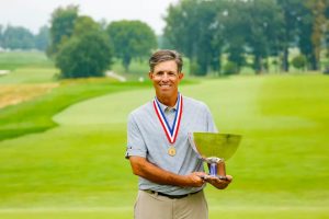 Man in patterned polo with gold medal and red-white-blue ribbon holding a silver trophy on a golf course fairway