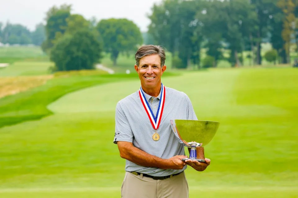 Man in patterned polo with gold medal and red-white-blue ribbon holding a silver trophy on a golf course fairway