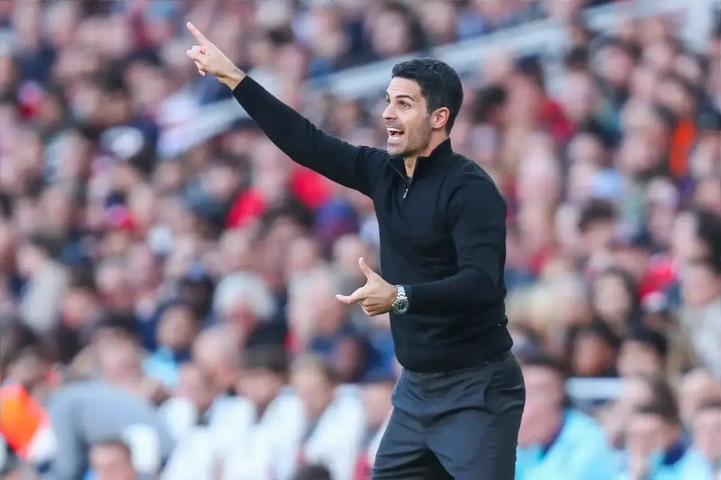 Man in black zip-neck top pointing and gesturing emphatically at a packed stadium crowd during a match