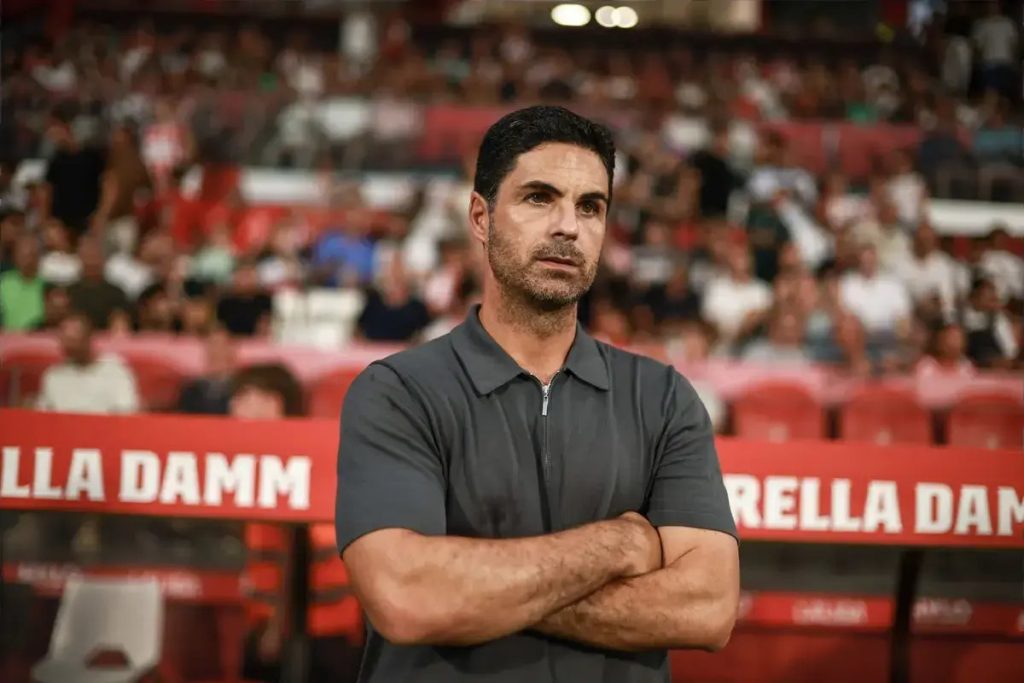 Mikel Arteta with arms folded in a dark polo shirt at a stadium, crowd and red advertising boards behind him.