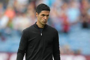 Mikel Arteta in a black zip-up top at a stadium, looking serious, with blurred fans behind him.