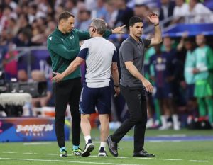 Man in a grey polo gesturing to the crowd on a football pitch, while a man in a green jacket and a staff member in a grey shirt stand beside him near a "Cham…