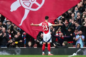 Mikel Merino celebrates in Arsenal's red and white kit, number 23, arms outstretched before a large flag and cheering fans at the stadium.