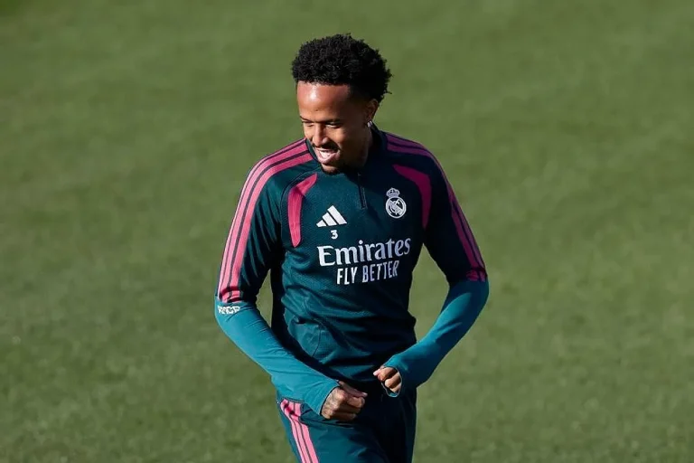 Jogador do Real Madrid, de sweat verde-escuro com detalhes rosa e número 3, sorri durante um treino num relvado.