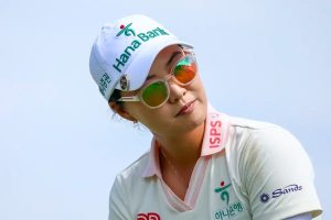 A female golfer in a white Hana Bank cap and white ISPS shirt with pink trim, wearing mirrored sunglasses and tilting her head against a pale sky.