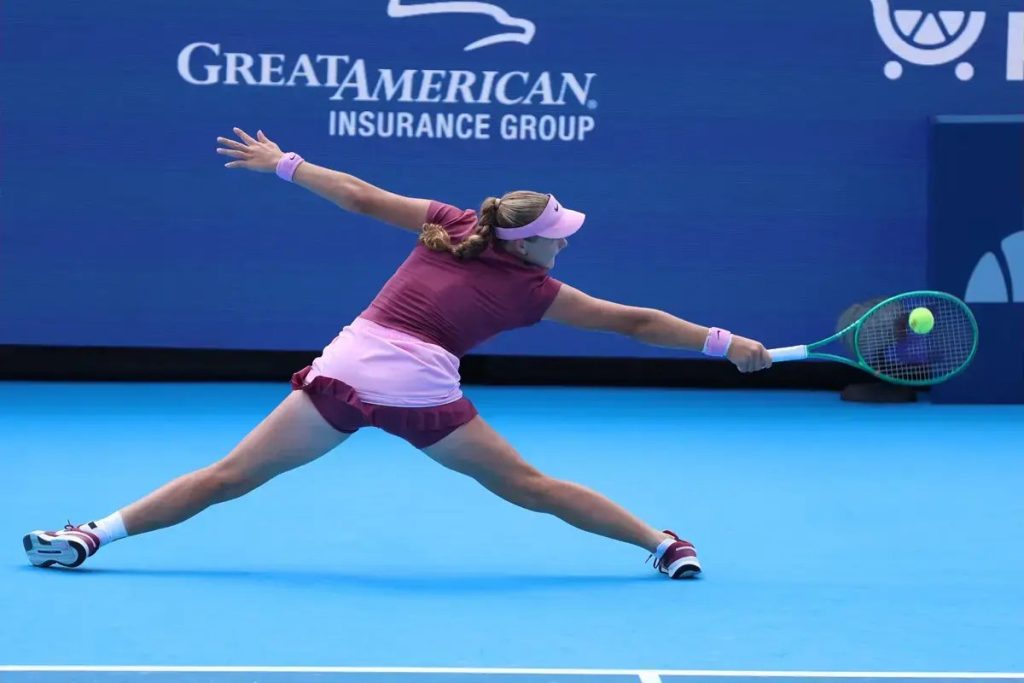 Tennis player in maroon and pink outfit and visor, stretching wide to hit a backhand on a blue court under a Great American Insurance Group banner.