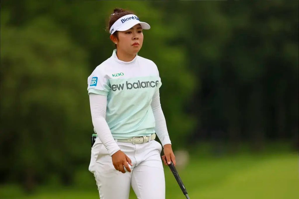 Golfer in mint green and white New Balance top and white visor with Biore logo, standing on a course with trees behind, holding a club