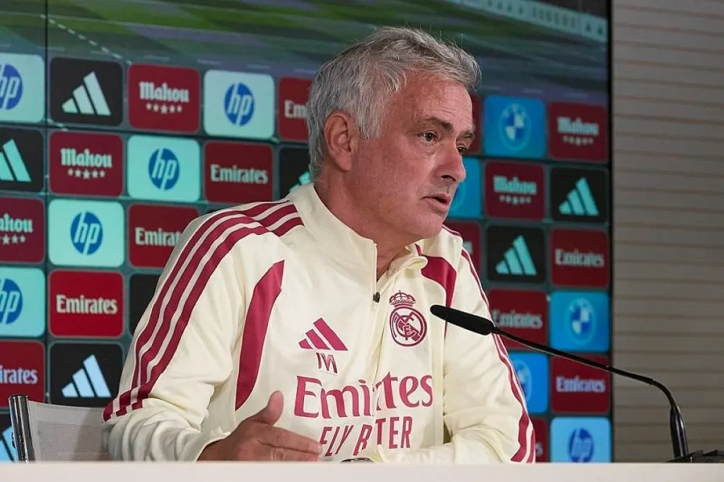 Treinador de cabelo grisalho com casaco Real Madrid em conferência de imprensa, diante de patrocinadores Emirates, HP e Mahou