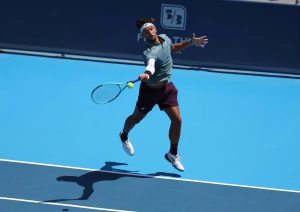 Tennis player in grey shirt and maroon shorts lunges to hit a forehand on a blue hard court, racket extended, ball in view.