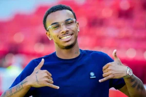 A smiling young man in glasses and a navy t-shirt making hand gestures at a stadium with red seats; he has tattoos on his arm and a watch, no visible name or…