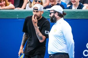 Man in black tee and white cap holds a racket beside an official in white shirt and cap, on a blue tennis court with spectators behind