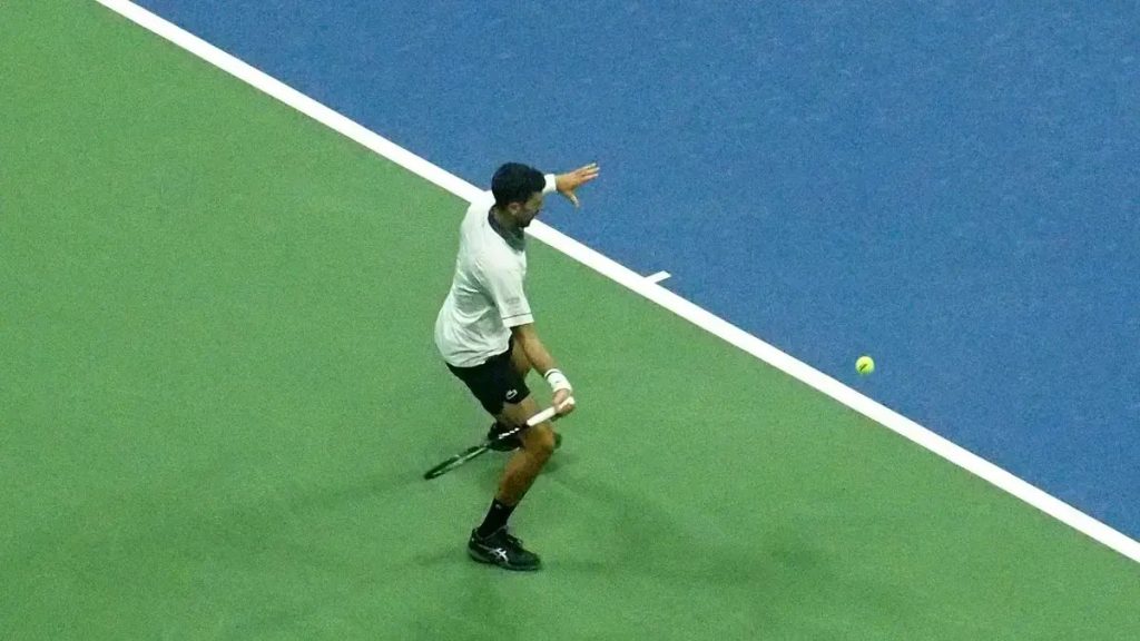 Tennis player in white shirt and black shorts hitting a low forehand on a blue and green court, ball in the air beside him