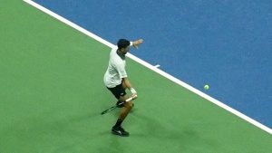 Tennis player in white shirt and black shorts hitting a low forehand on a blue and green court, ball in the air beside him