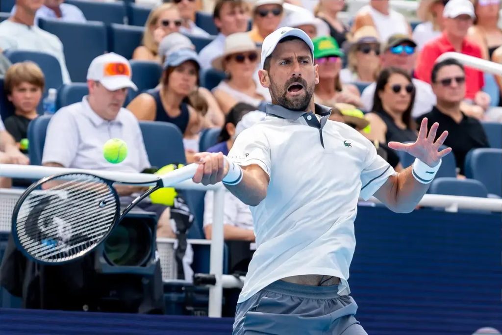Tennis player in a white Lacoste polo and white cap, mid-stroke with a racket, mouth open, arm outstretched, while spectators watch courtside.