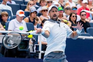 Tennis player in a white Lacoste polo and white cap, mid-stroke with a racket, mouth open, arm outstretched, while spectators watch courtside.