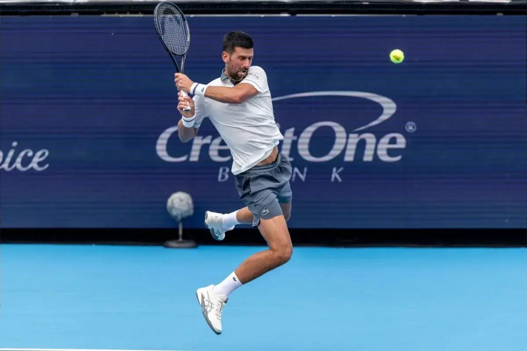 Tennis player in white shirt and grey shorts hits a forehand mid-jump on a blue court in front of a CreditOne Bank sign