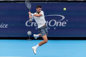 Tennis player in white shirt and grey shorts hits a forehand mid-jump on a blue court in front of a CreditOne Bank sign