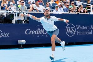 Tennis player in white polo and grey shorts lunges full stretch to reach a ball on a blue court, sponsor boards behind him.