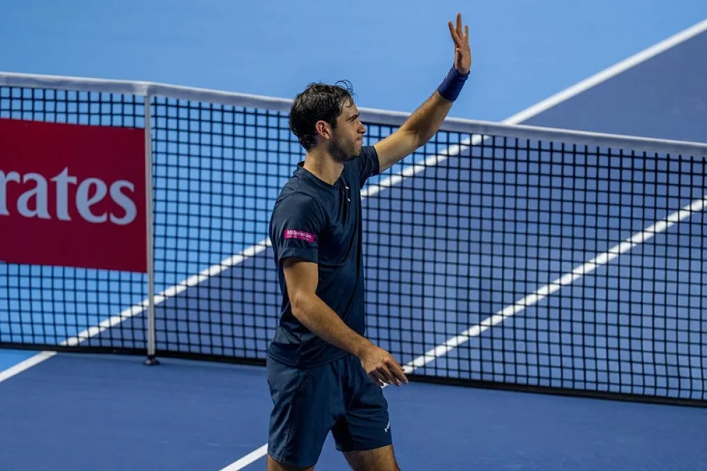 Homem de ténis de camisola azul-marinho acena com a mão levantada junto à rede, num court azul, com patrocínio Emirates.