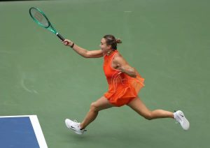 Tennis player in an orange sleeveless dress and white sneakers stretching for a forehand with a green racket on a green hard court at the US Open