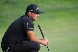 Golfer in black cap and shirt crouching on a green, gripping a putter and studying his line. Caps read "Grindworks" and "A" logos.
