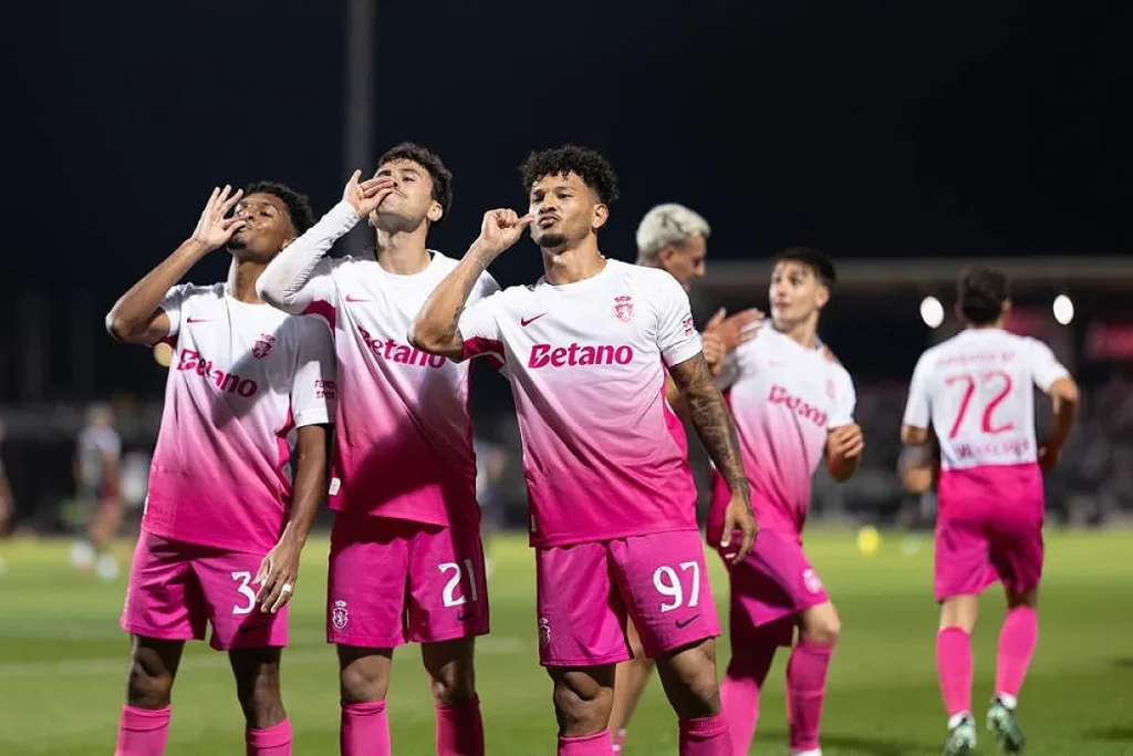Jogadores do Casa Pia com camisola cor-de-rosa e Betano celebram um golo à noite, com o número 97 em destaque.
