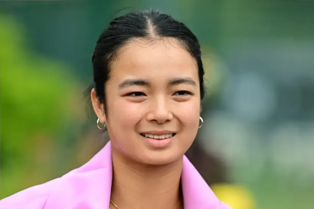 Woman with dark hair pulled back, gold hoop earrings and a pink blazer, smiling outdoors against a blurred green background, tennis setting