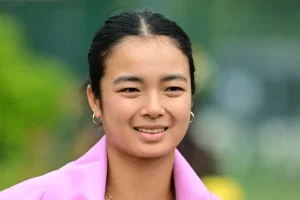 Woman with dark hair pulled back, gold hoop earrings and a pink blazer, smiling outdoors against a blurred green background, tennis setting