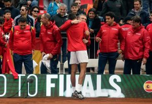 Players in red Spain team jackets at a clay court; a player in red shirt embraces a teammate near a "To Finals" banner with Spanish flag