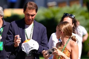 A man in a navy blazer and white cap signing a Boss cap for a young woman holding a phone, in a paddock setting