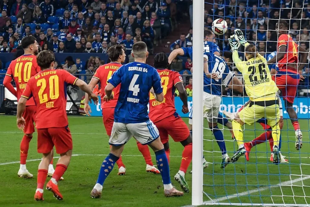 Jogadores do RB Leipzig de vermelho e do Schalke 04 de azul disputam uma bola na área, com o guarda-redes de amarelo n.º 26 a defender.